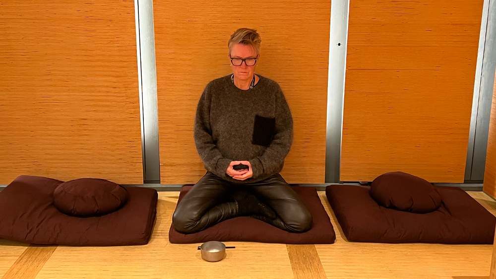 Kvinne sitter i lotusstilling på mørke meditasjonsmatter. Hun folder hendene og ser ned på en syngebolle.
