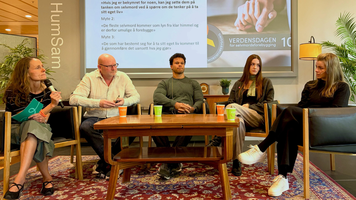 Five people sit around a table on a stage. Behind them is a screen with the logo for World Suicide Prevention Day.